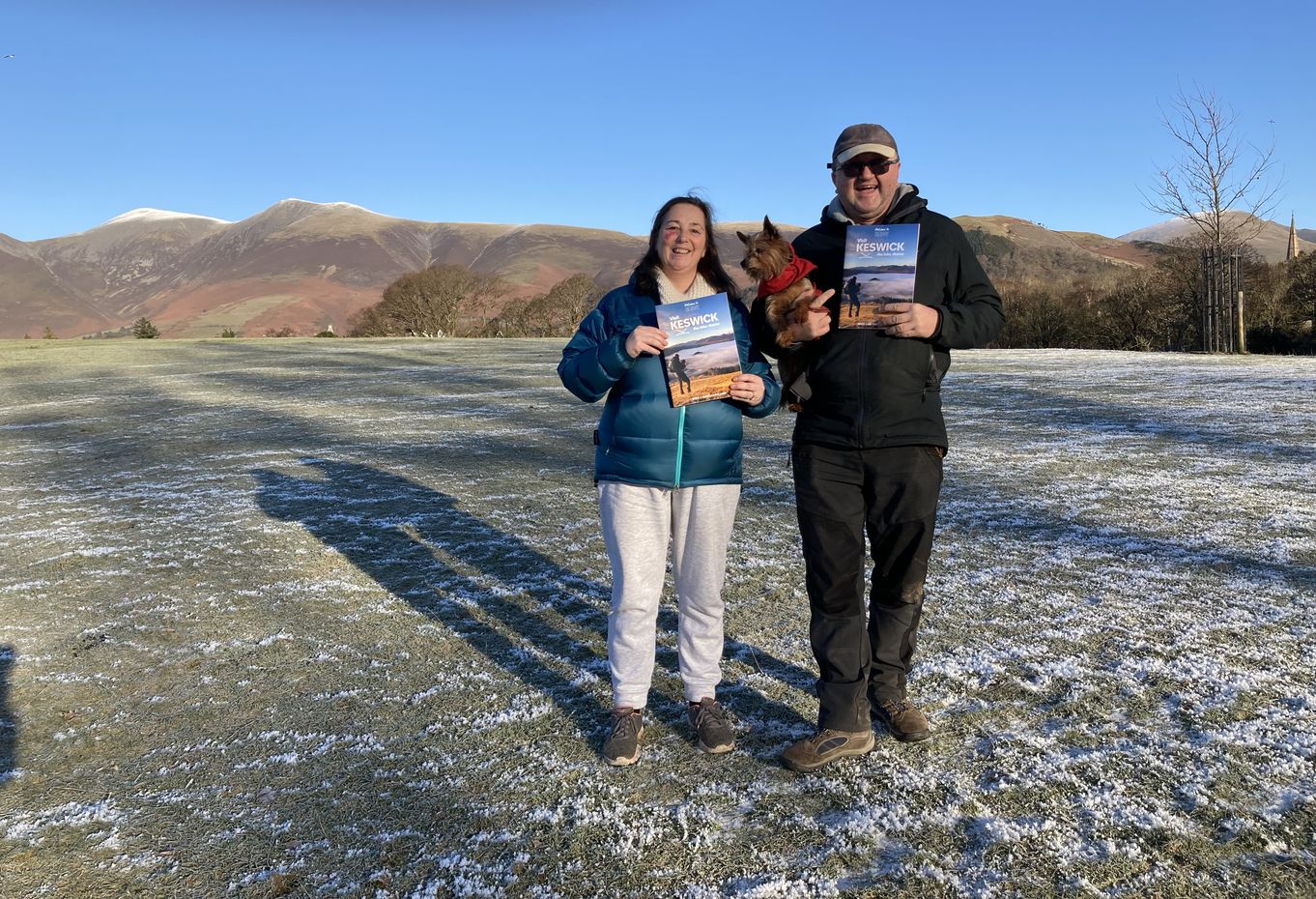 Vanessa Metcalfe and Rob Knowles standing in Crow Park with Skiddaw and Latrigg behind holding the Visit Keswick Guide.  Robin is also holding his dog, Rosie